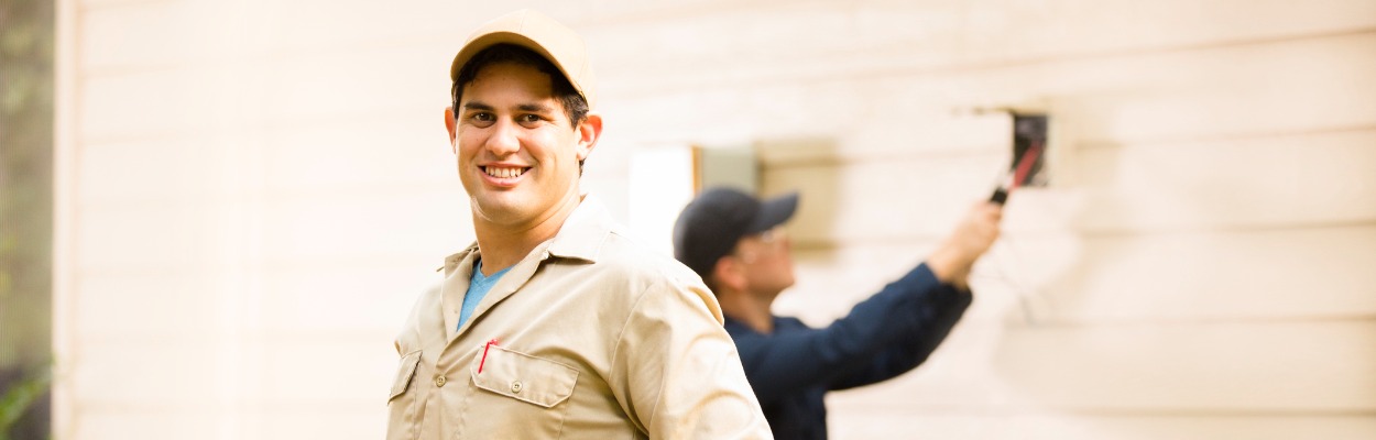 An Hvac Tech Smiling For The Camera While Another Works Behind Him